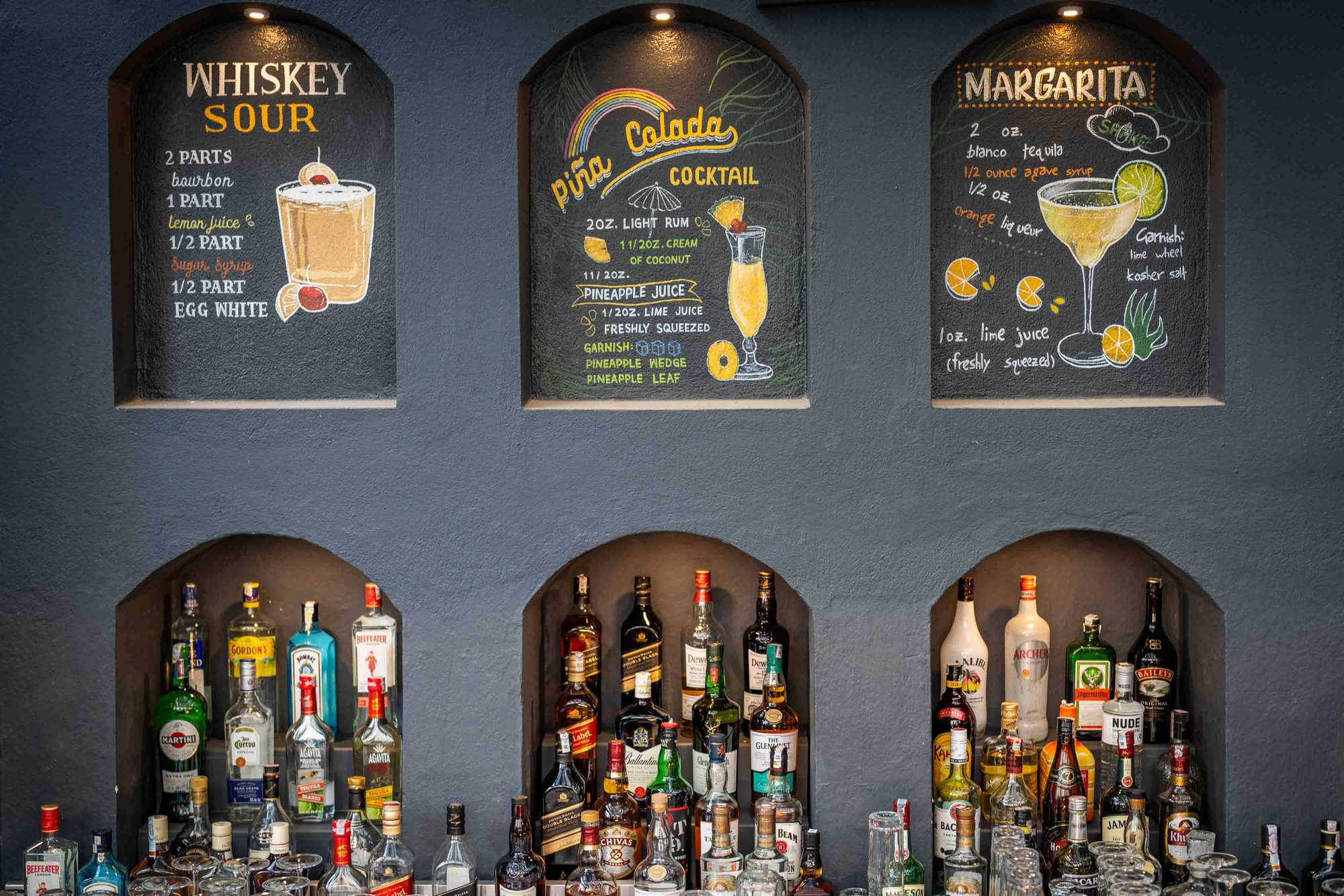 Hand-chalked whiskey sour, pi&ntilde;a colada, and margarita recipes above the lit spirits niches at Jharana Bar